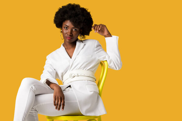 beautiful african american woman sitting on yellow chair isolated on orange 