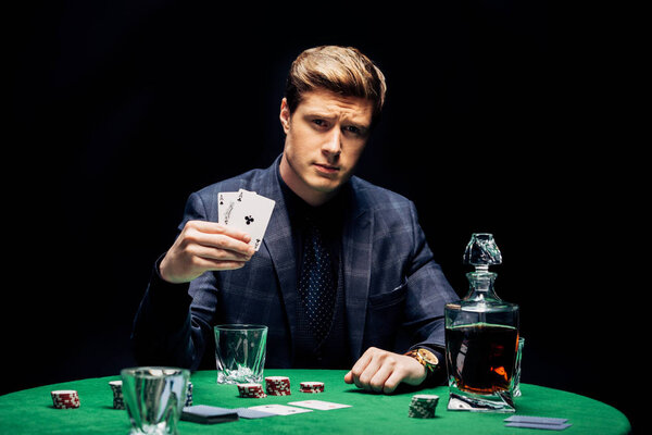 KYIV, UKRAINE - AUGUST 20, 2019: selective focus of handsome man holding playing cards near poker chips and alcohol drink isolated on black 
