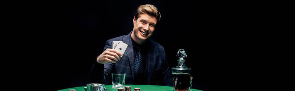 KYIV, UKRAINE - AUGUST 20, 2019: panoramic shot of happy man holding playing cards near alcohol drink isolated on black 