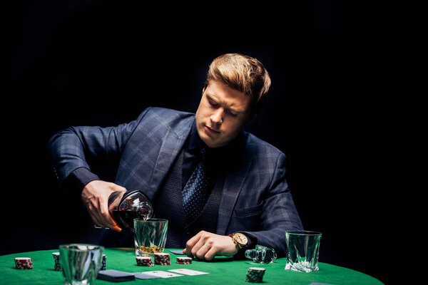 KYIV, UKRAINE - AUGUST 20, 2019: selective focus of handsome man pouring whiskey in glass near playing cards isolated on black 