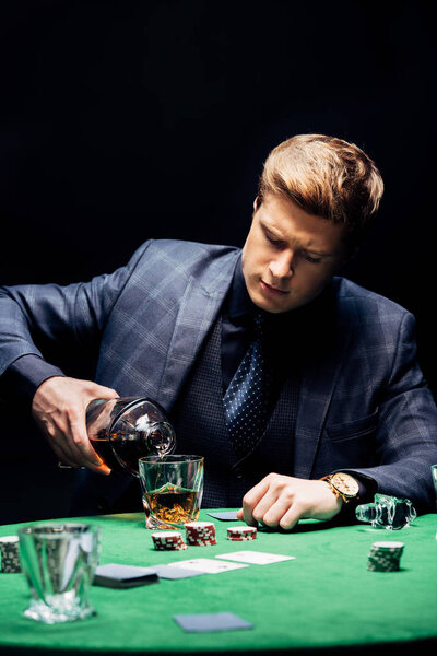 KYIV, UKRAINE - AUGUST 20, 2019: selective focus of man pouring whiskey in glass near playing cards isolated on black 
