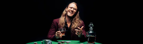 全景拍摄的快乐胡子男子戴着眼镜拿着玻璃与威士忌附近的扑克牌隔离在黑色 — 图库照片