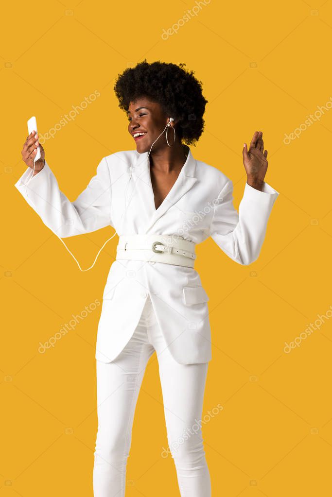 cheerful african american girl listening music while holding smartphone and gesturing isolated on orange 