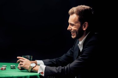 happy bearded man smiling near poker table isolated on black  clipart
