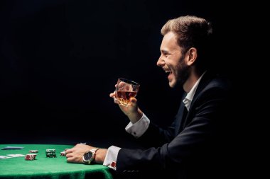 mutlu sakallı adam gülümseyen ve siyah izole poker masası yakınında cam tutan yan görünümü 