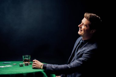 cheerful man smiling near poker table with glass of whiskey on black  clipart