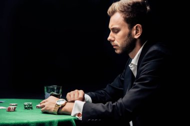 side view of upset man playing poker near glass of whiskey isolated on black  clipart