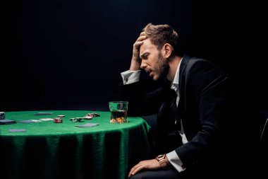 KYIV, UKRAINE - AUGUST 20, 2019: side view of sad man playing poker near glass of whiskey isolated on black  clipart