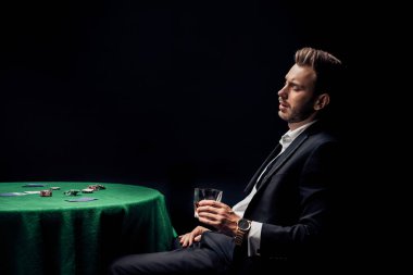 upset bearded man holding glass near poker table isolated on black  clipart