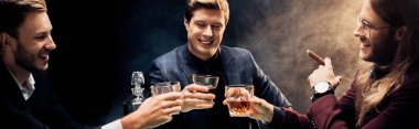panoramic shot of happy men clinking glasses with whiskey on black with smoke clipart