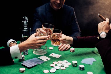 KYIV, UKRAINE - AUGUST 20, 2019: cropped view of men toasting glasses near poker table on black with smoke  clipart