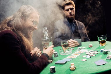 Kyiv, Ukrayna - 20 Ağustos 2019: Poker masasının yanında poker fişleri ve dumanla kağıt oynayan yakışıklı adamlar 