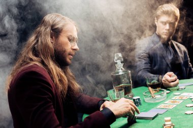 Kyiv, Ukrayna - 20 Ağustos 2019: Poker masasının yanında poker oynayan yakışıklı adamlar ve siyah üzerinde dumanla kağıt oynuyorlar 