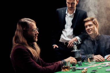 KYIV, UKRAINE - AUGUST 20, 2019: handsome friends gesturing while playing poker on black with smoke  clipart