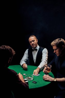 Kyiv, Ukrayna - 20 Ağustos 2019: Sakallı krupiyenin seçici odak noktası poker masasına siyah üzerine siyah dumanlı adamların yanına oyun kartları koymak 