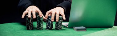 KYIV, UKRAINE - AUGUST 20, 2019: panoramic shot of  man touching poker chips near laptop isolated on black clipart
