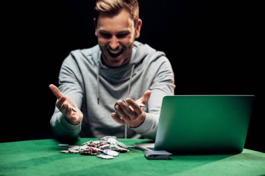KYIV, UKRAINE - AUGUST 20, 2019: selective focus of happy man looking at poker chips near laptop isolated on black  clipart