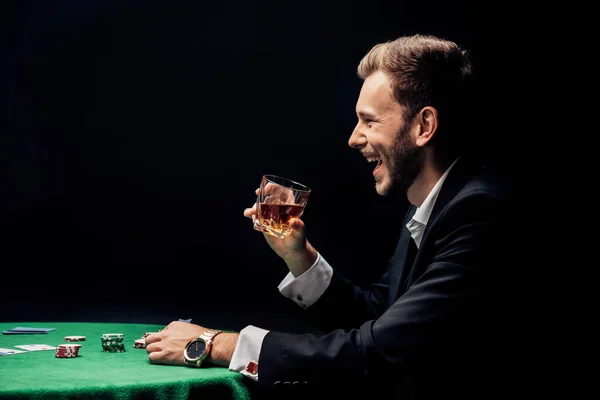 侧视图的快乐胡子男子微笑和持有玻璃附近的扑克桌孤立黑色 — 图库照片