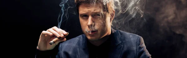 全景拍摄的英俊男子吸烟雪茄在黑色与烟雾 — 图库照片