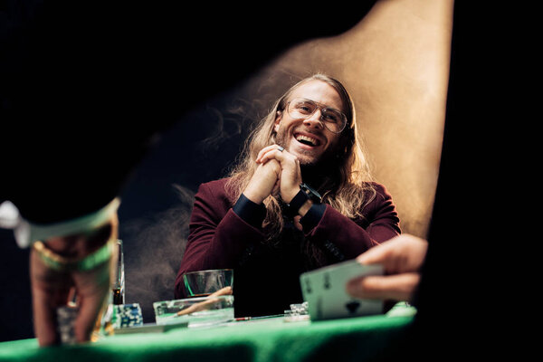 KYIV, UKRAINE - AUGUST 20, 2019: selective focus of happy bearded man in eye glasses playing poker with friend on black with smoke 