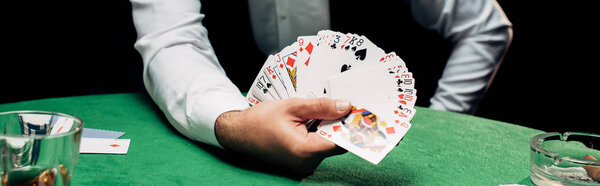КИЕВ, УКРАИНА - 20 августа 2019 года: панорамный снимок крупье в формальной одежде, держащего игральные карты возле покерного стола, изолированного на черном

