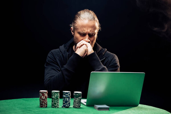 KYIV, UKRAINE - AUGUST 20, 2019: man covering face while using laptop near poker chips and playing cards on black with smoke