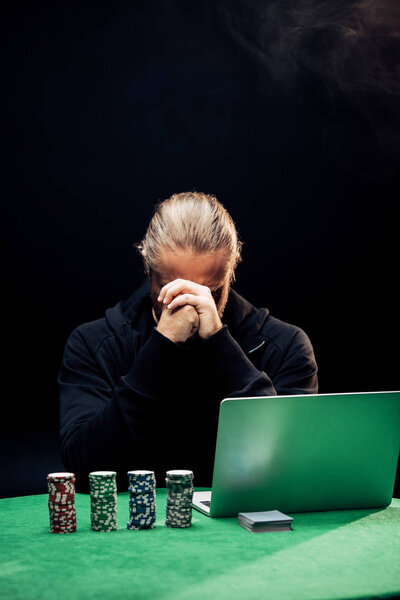 KYIV, UKRAINE - AUGUST 20, 2019: man covering face while using laptop near poker chips on black with smoke