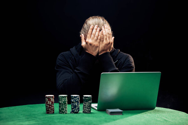 KYIV, UKRAINE - AUGUST 20, 2019: man covering face while using laptop near poker chips isolated on black 