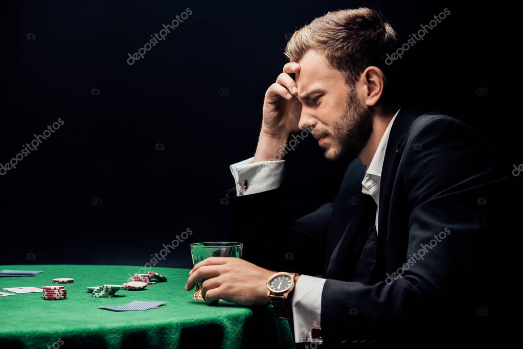 Frustrated man holding glass near poker table isolated on black