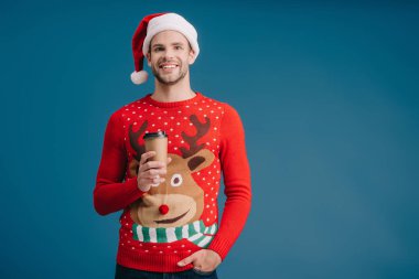 Gülümseyen adam Noel Baba şapkası ve Noel süveteri içinde kahve tutuyor.