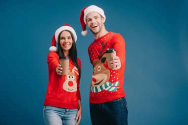 Noel Baba şapkalı ve Noel süveterli neşeli bir çift mavi üzerinde izole olmak için kahve tutuyorlar.