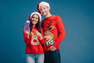 Kyiv, Ukrayna - 06 Ağustos 2019: Noel Baba şapkalı ve Noel kazaklı çift Instagram uygulamalı akıllı telefona bakıyor, mavi üzerine izole edilmiş.