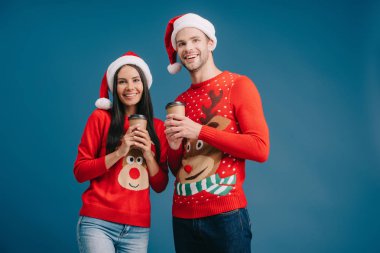 Noel Baba şapkalı ve Noel süveterli gülümseyen bir çift mavi üzerinde izole olmak için ellerinde kahve tutuyorlar.