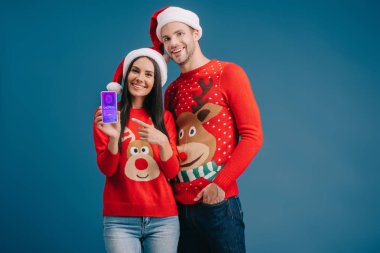 Noel Baba şapkalı ve Noel süveterli mutlu çift akıllı telefona alışveriş uygulaması ile bakıyor, mavi üzerine izole edilmiş.