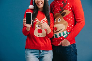 Noel Baba şapkalı ve Noel süveterli çift mavi ekranda boş ekranlı akıllı telefon gösteriyor.