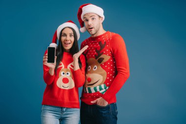 Noel Baba şapkalı ve Noel süveterli sürpriz çift mavi ekranda boş ekranlı akıllı telefon gösteriyor.