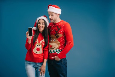 Noel Baba şapkalı ve Noel süveterli mutlu çift mavi ekranda boş ekranlı akıllı telefon gösteriyor.