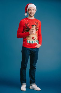 Noel Baba şapkası ve Noel süveteri giymiş yakışıklı, mutlu bir adam elinde kahve tutuyor.