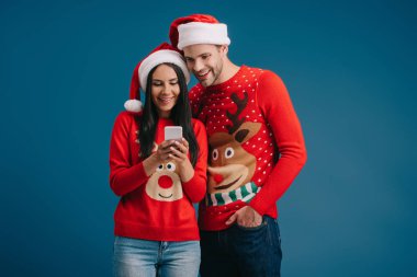 Noel Baba şapkalı ve kışlık kazaklı çift mavi üzerine izole edilmiş akıllı telefon kullanıyor.