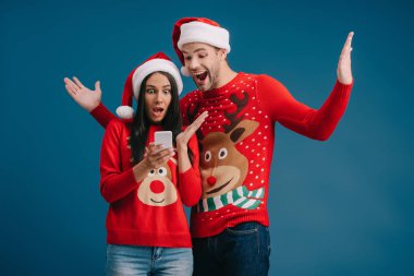 Noel Baba şapkalı ve Noel süveterli heyecanlı çift mavi üzerine izole akıllı telefon kullanıyor.