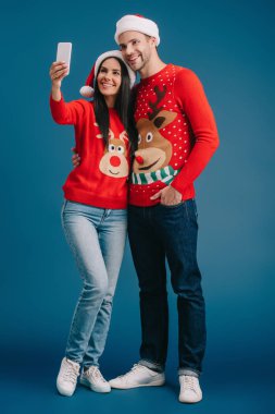 santa şapka ve noel kazak mavi izole akıllı telefon selfie alarak gülümseyen çift