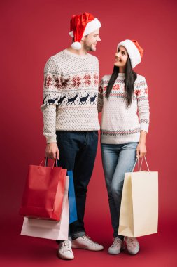 santa şapkalı mutlu çift birbirlerine bakıyor ve kırmızı izole alışveriş çantaları tutarak