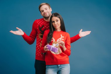 girlfriend holding christmas present, boyfriend standing behind with shrug gesture isolated on blue clipart