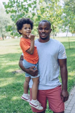 Mutlu Afro-Amerikalı adam sevimli oğlunu tutuyor ve parkta yürürken kameraya bakıyor. 