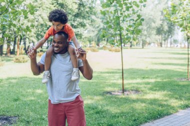 Yakışıklı Afrikalı Amerikalı adam parkta yürürken sevimli oğlunu sırtında taşıyor.