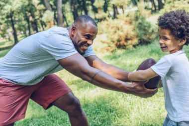 Neşeli Afrikalı Amerikalı adam parkta sevimli oğluyla rugby oynuyor.