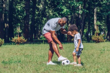 Genç Afrikalı Amerikalı adam parkta futbol oynayan sevimli oğlanı eğitiyor.