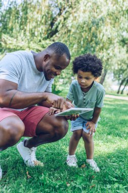 Gülümseyen Afrikalı Amerikalı adam parkta yakışıklı oğluna dijital tablet gösteriyor.