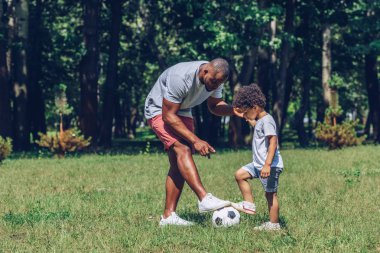 Genç Afrikalı Amerikalı baba parkta oynayan oğluna futbol öğretiyor.