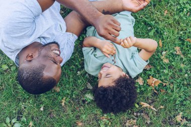 overhead view of african american father and son having fun while lying on lawn in park clipart